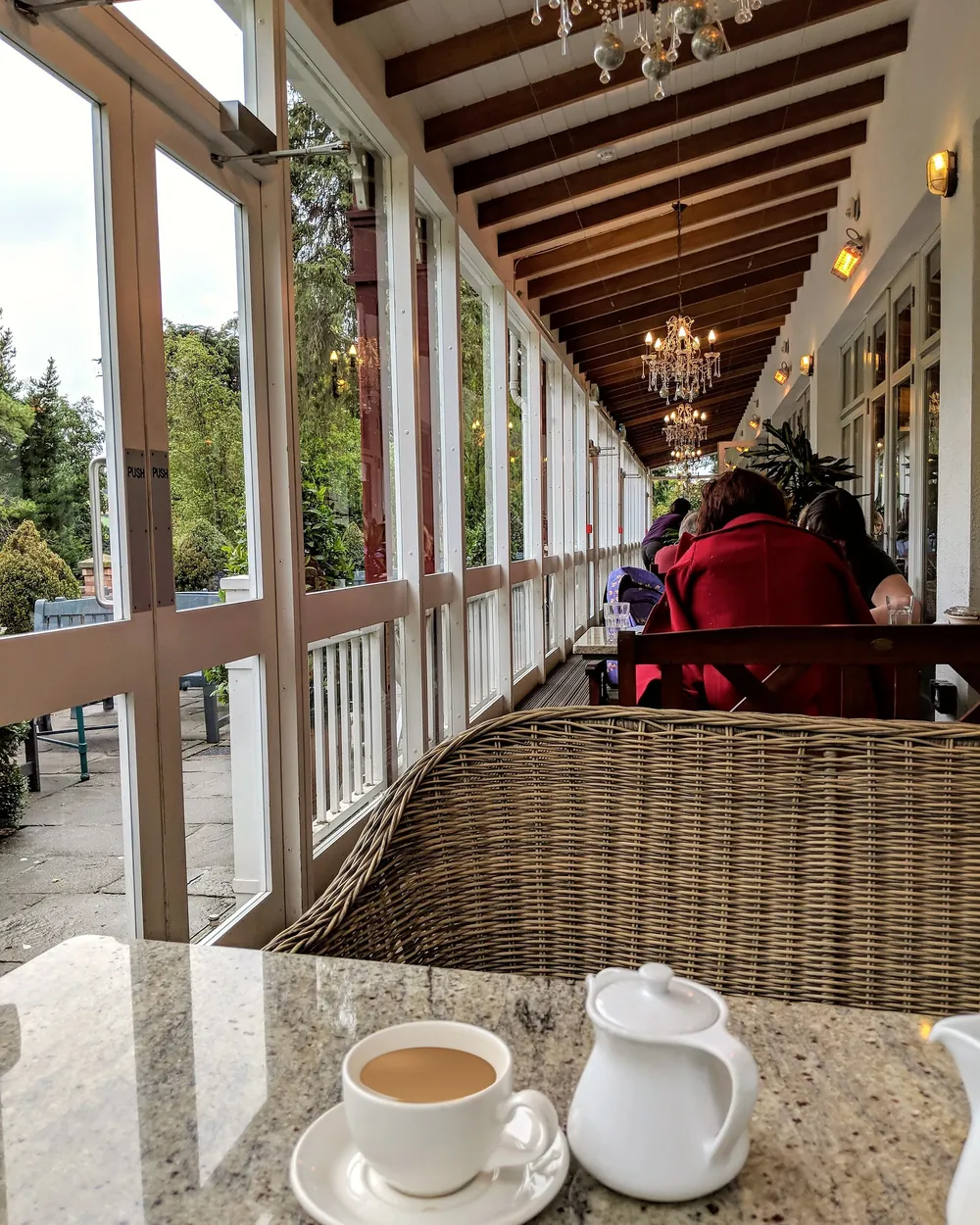 A cup of tea and a milk jug on a stone table in a long glass conservatory, with wicker chairs and chandeliers