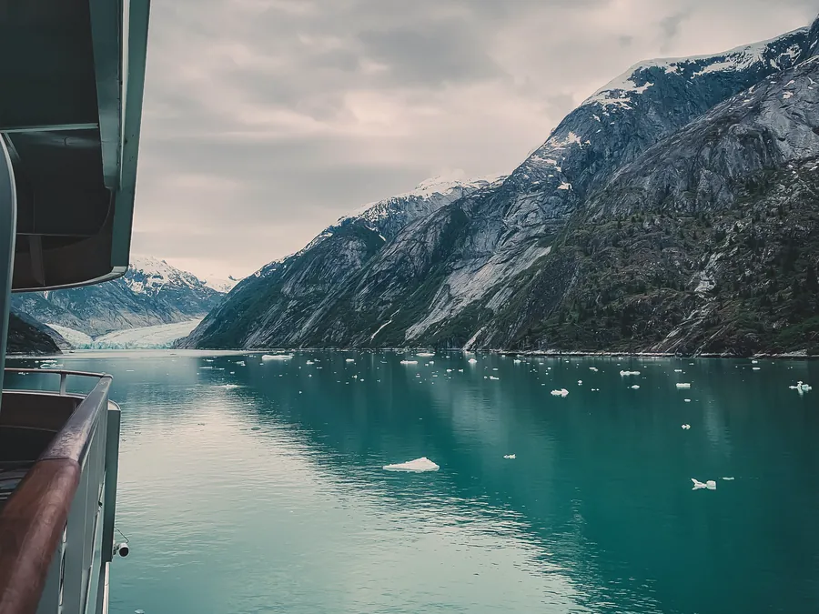 Calm green water with floating ice and snowy mountains, seen from the side of a ship