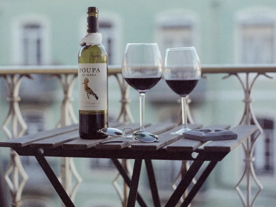 Two glasses of red wine, a bottle and a pair of sunglasses on a small table on a balcony with an ornate railing
