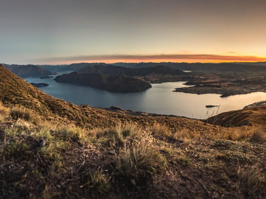 A grassy hilltop trail above a winding lake, with the sky glowing gold on the horizon
