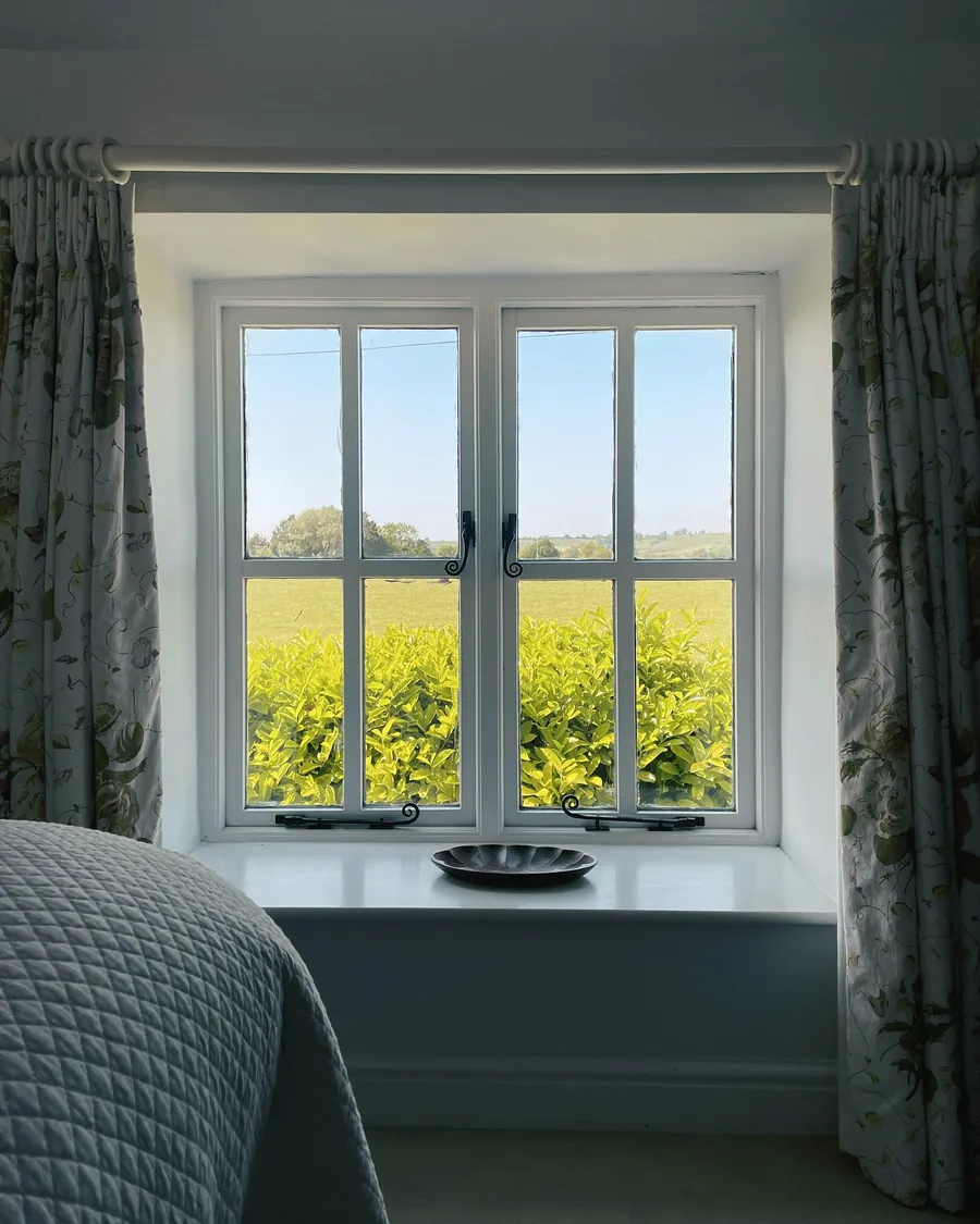 A bedroom window with floral curtains looking out over green and yellow fields