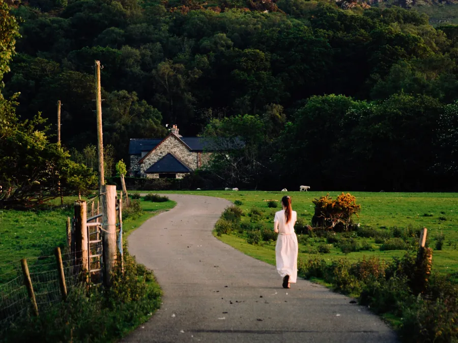 A woman walking up a quiet lane toward a cottage below a mountain