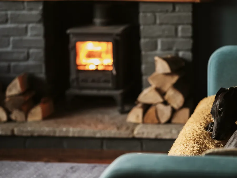A wood stove glowing beside stacked logs, seen past the arm of a sofa